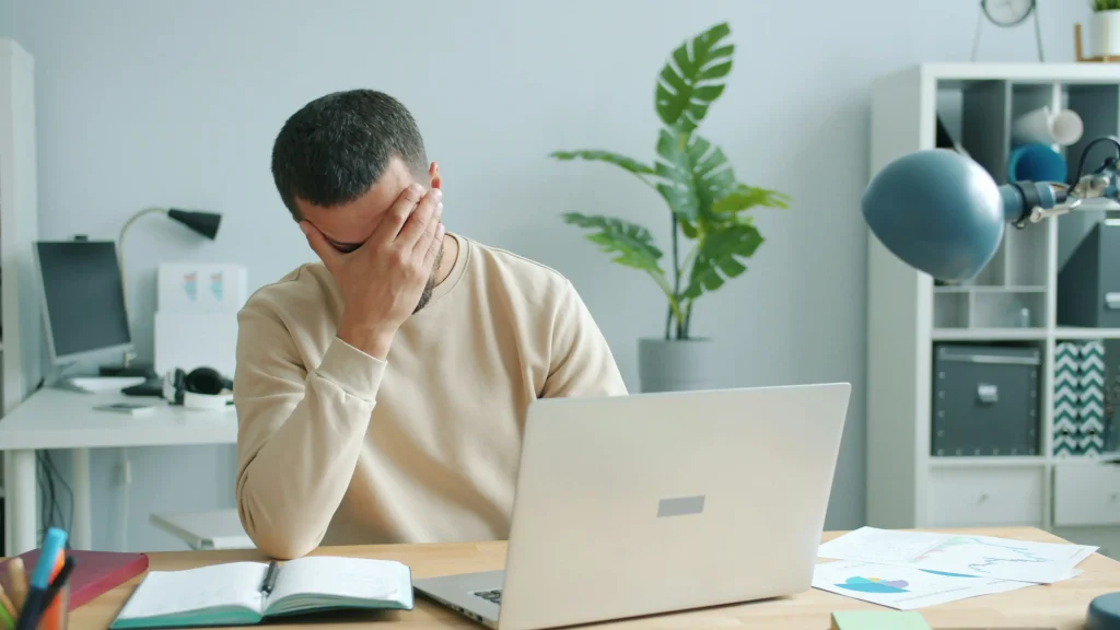 Overworked in-house employee facepalming at a chaotic desk, representing high workload and business inefficiency compared to hiring virtual assistants